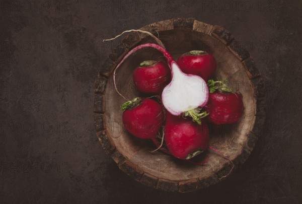 A collection of vibrant red radishes sits in a carved wooden bowl on a dark, textured surface. The