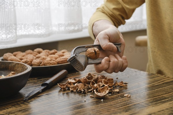 A person is breaking open a walnut with a metal nutcracker, sitting at a wooden table. Shelled nuts