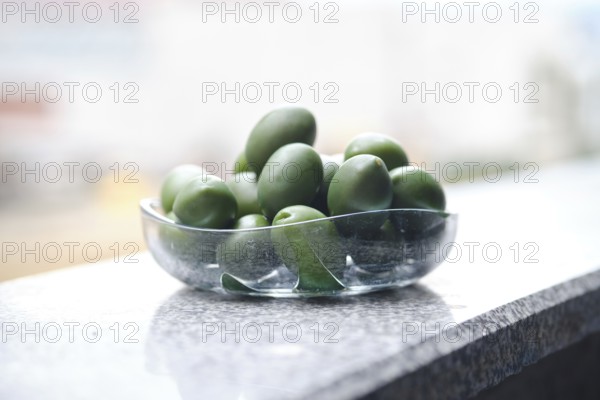 A bowl filled with green olives on a smooth granite surface. The sunlight reflects off the glass