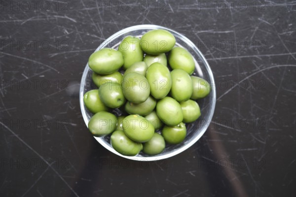 Overhead view of a bowl filled with green olives on a black surface. The olives are vibrant and