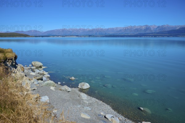 Rocky shore and clear lake with mountain views under clear skies, Lake Pukaki Viewpoint, Pukaki,