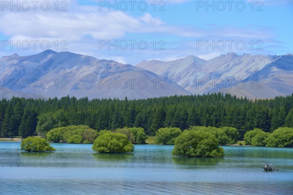 A lake with small green islands and wooded slopes against a mountain range and blue sky, Lake