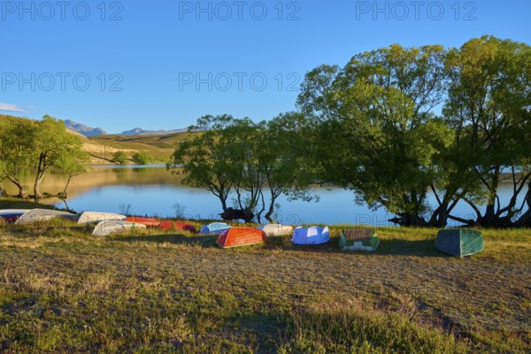 Boats are tipped over on the shores of a calm lake in the evening with wooded hills, Lake McGregor,
