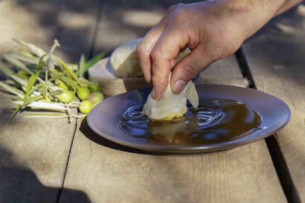 Hand dipping a piece of fresh bread into a shallow plate of extra virgin olive oil, with olives and