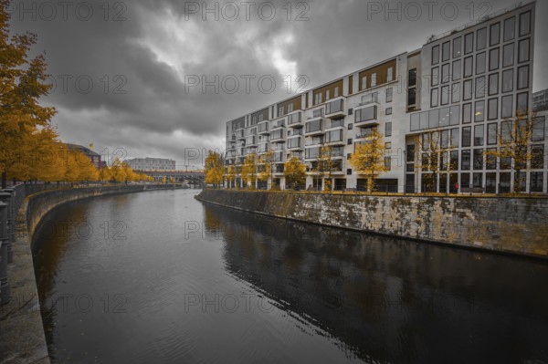 Modern apartment block on the river with yellow autumn trees, dramatic sky reflected in calm water,