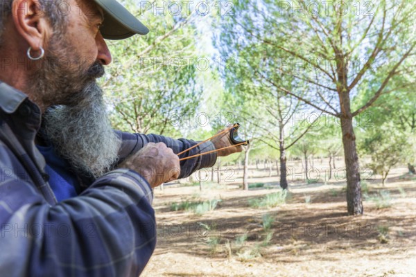 Man with a long beard holding a slingshot, aiming a projectile in a forest environment. Concept of
