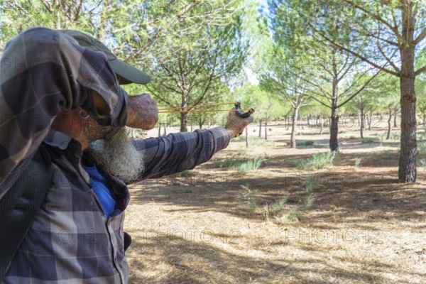 Man with beard and plaid shirt aiming a slingshot, focusing intently on a target in a pine forest