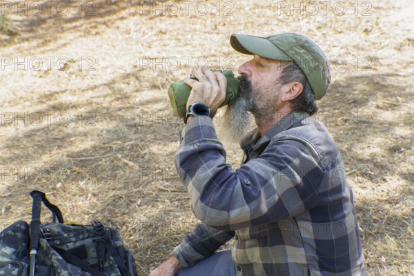 Mature bearded hiker drinking from a canteen during a trekking break in sunlit forest, resting and