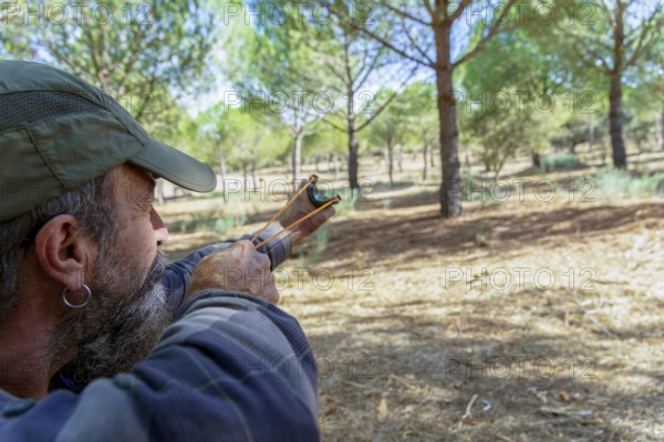 Bearded man in cap aims a slingshot in a sunlit pine forest, focused on target during outdoor