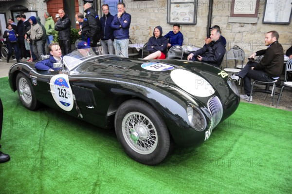 Black jaguar roadster rolls across green carpet during a historic rally, spectators sitting close