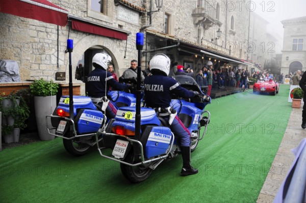 Police motorcycles escort a classic car rally through the stone old town, audience lines the green