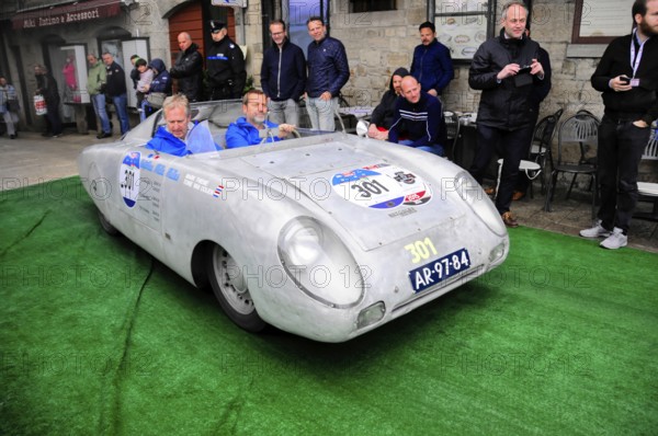 Aluminum-colored racing roadster with blue drivers at a vintage car rally on green carpet, purist