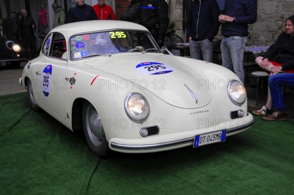 White Porsche 356 with glowing headlights drives across green carpet during a vintage car rally,