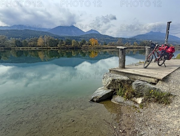 An idyllic lake with a bicycle on a jetty, surrounded by autumnal landscape and mountains, Drau,