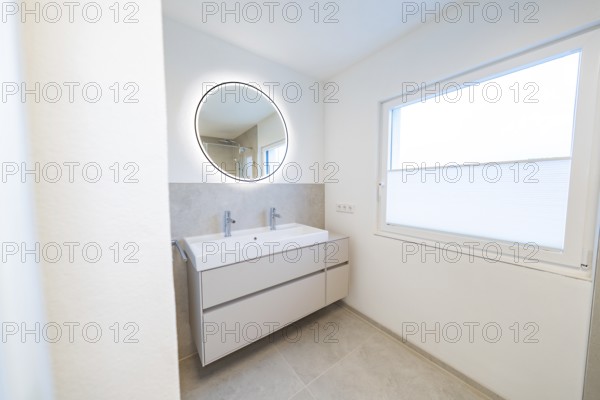 Bright bathroom with round illuminated mirror and double washbasin in front of subtle shades of