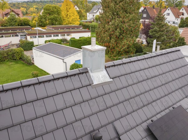Roof with anthracite-colored tiles and modern chimney, behind it quiet residential area with autumn