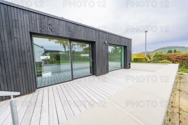 Large terrace with light wooden deck against black façade and sliding doors, wide views of autumn