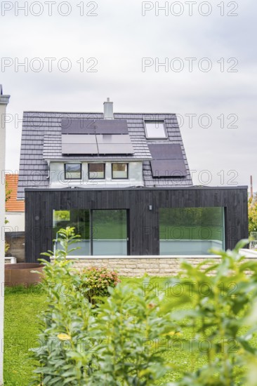 Front view of a house with solar panels and black extension with glass front, well-kept garden and