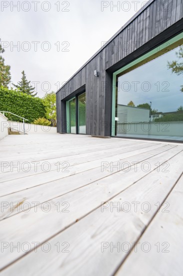Deep view along the wide wooden terrace of the modern, black-clad building with floor-to-ceiling