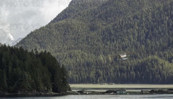 A seaplane flies over a tranquil lake surrounded by dense forest and mountains under an overcast