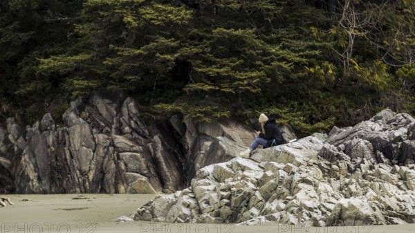 A person sits on rocky coastline surrounded by trees under a cloudy sky in Tofino, British