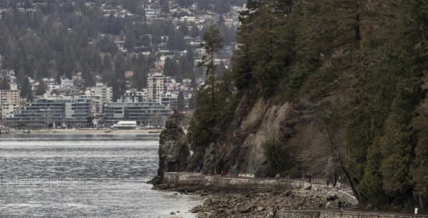Rocks and trees line a coastal path in Stanley Park with urban buildings in the distance in