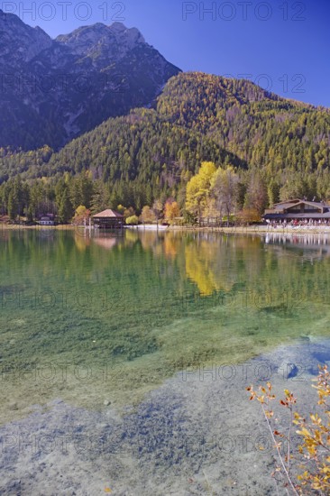 Emerald green, clear shore water with visible stones, colorful autumn trees and a boathouse are
