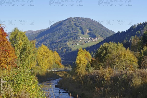 A stream snakes through an autumnal valley, on the sunny mountainside there is a village under deep