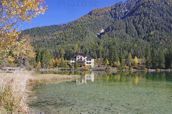 Clear shore water of an alpine lake, opposite is a villa reflected against colorful autumn forests