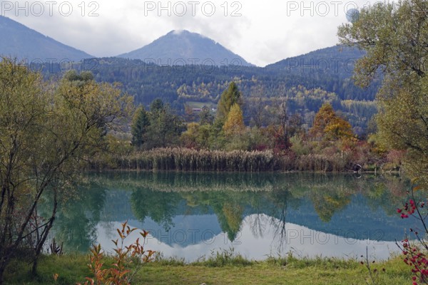 Quiet lake with turquoise-green water, autumnal shore and mountain scenery under cloudy sky, Drau,
