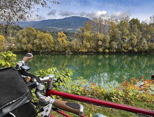 View over the handlebar of a shimmering green river in front of autumn trees and mountains, bicycle