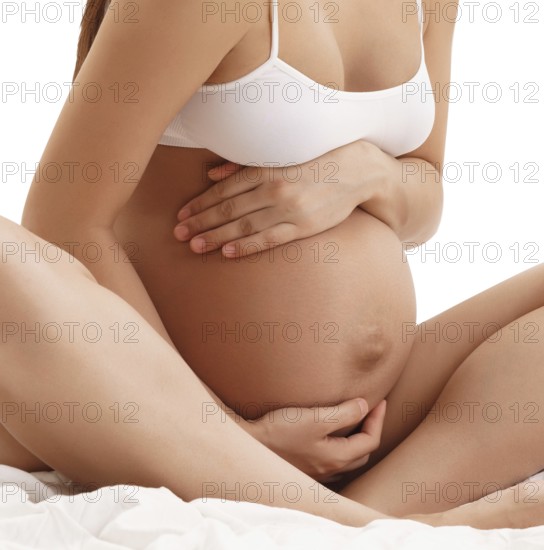 Young long haired pregnant brunette in white top sitting on bed posing holding her hands on her