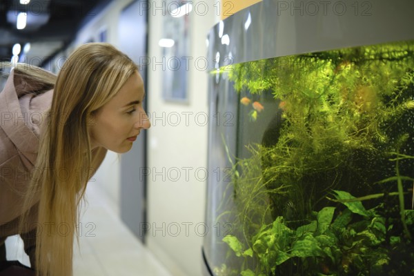 A woman with long hair leans forward, peering into a large aquarium filled with vibrant fish and