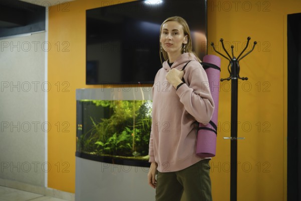 A woman in a pink hoodie stands in a fitness studio, holding a yoga mat. An aquarium with aquatic