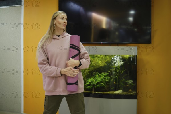 A woman stands in a fitness studio, holding a yoga mat, looking inspired. An aquarium filled with