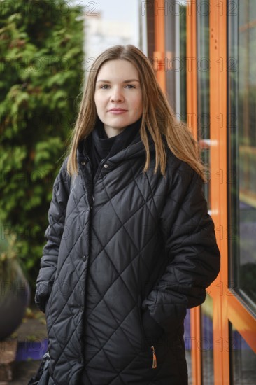 A young woman wearing a black quilted coat stands confidently outside a building. She has long hair