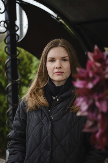 A young woman with long hair poses confidently near vibrant flowers in a garden. The scene captures