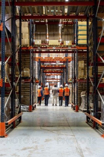 Workers in hard hats and reflective vests walk down a high rack warehouse aisle filled with stacked
