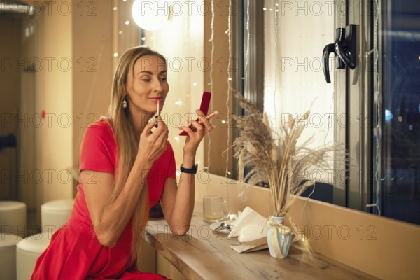 A woman dressed in a bright red dress applies lipstick in front of a mirror. She sits at a wooden