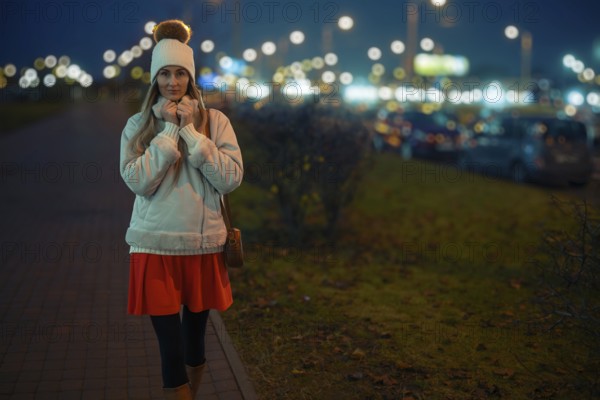 A woman walks on a city sidewalk at night, wearing a warm white jacket and a beanie. The street is