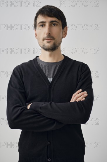 A man stands with arms crossed, portraying confidence. He wears a black cardigan over a gray shirt.