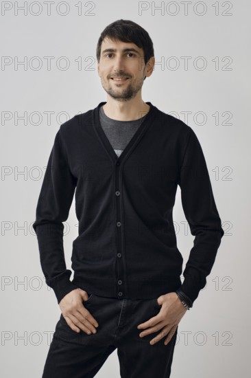 A man stands confidently with his hands on his hips, wearing a black cardigan over a t-shirt. The