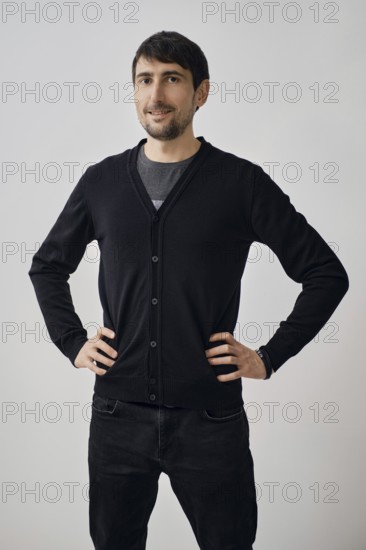A man poses confidently with his hands on his hips, wearing a black cardigan over a gray shirt. He