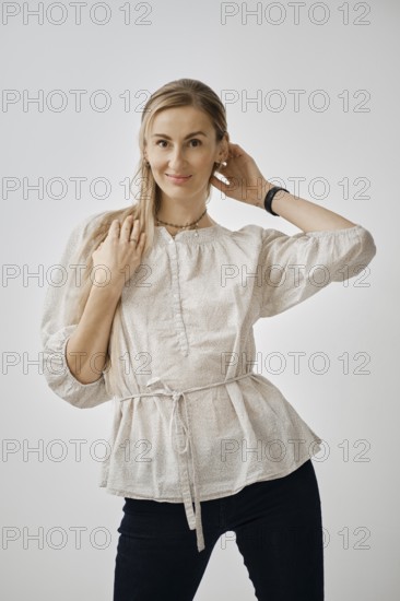 A playful woman stands in a bright studio, wearing a fashionable light blouse and dark pants. She