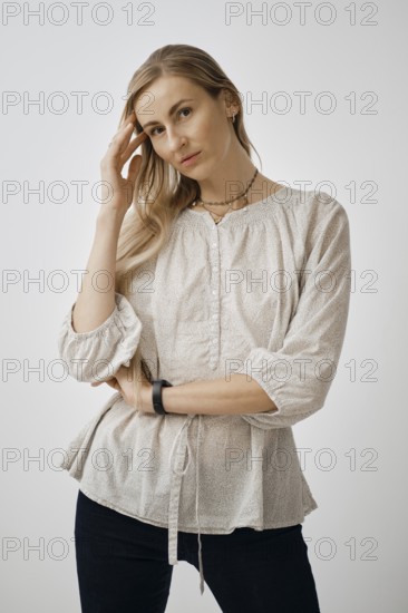 A pensive woman is standing with one hand touching her hair while wearing a light, textured blouse.