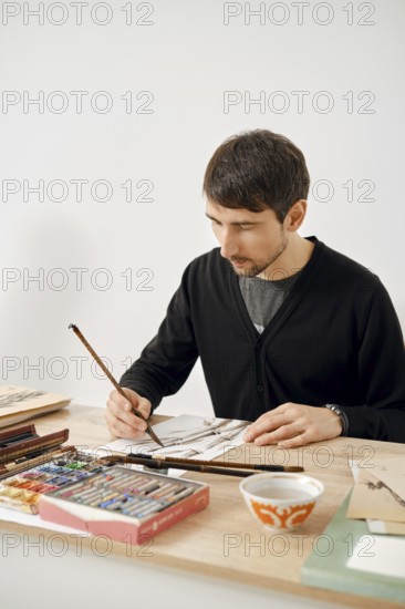 A male artist focuses on his work at a wooden table filled with various art supplies. He uses