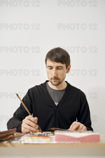 A focused artist is working diligently on a paper in a clean, bright studio. He uses a brush to add