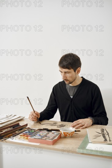 A focused artist sits at a wooden table, working on an intricate drawing with a brush