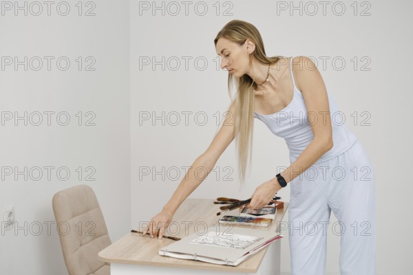 A woman dressed in light clothing is focused on arranging her art supplies and sketches on a table