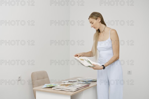 A woman with long hair stands in a bright, minimal room, looking through art sketches. She focuses
